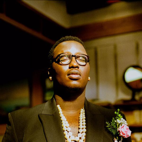 Artist SIPHO. wearing a black blazer with a bare chest and necklaces, holding their palms up in front of them and their eyes closed.