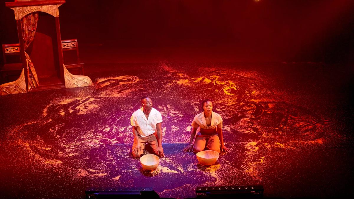 Two performers sit on a stage covered in sand. They have wooden bowls in front of them and a confession box structure stands behind them stage left. The stage is lit with red lights. 