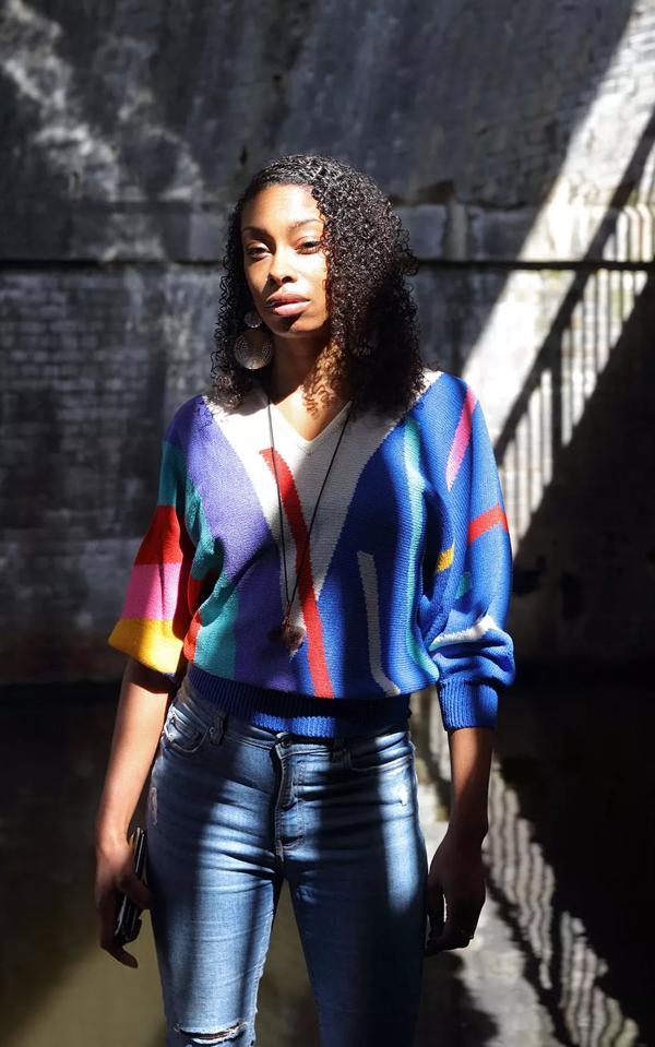 Ioney Smallhorne standing in front of a brick wall wearing a colourful patterned jumper and blue jeans.