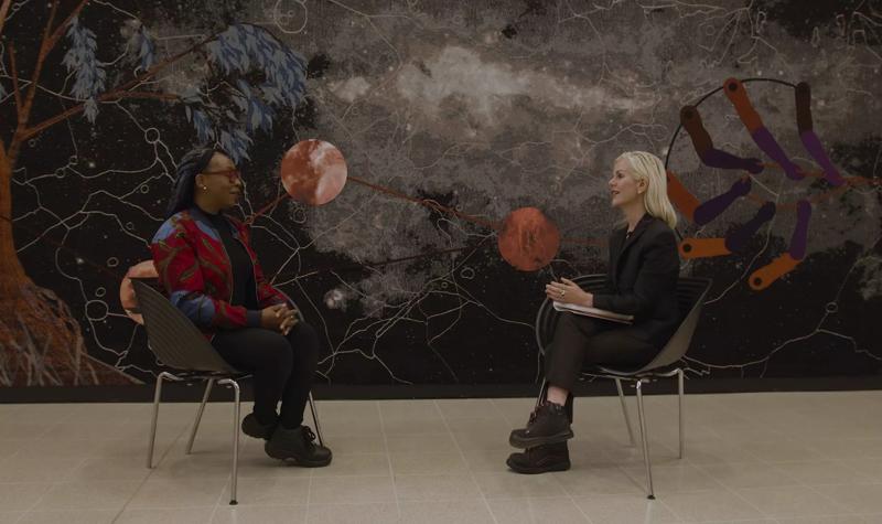 The artist Otobong Nkanga wearing red framed glasses and a multi-coloured jacket sits facing Rachel Thomas who wears all black. The pair are seated in front of one of Nkanga's large tapestry works