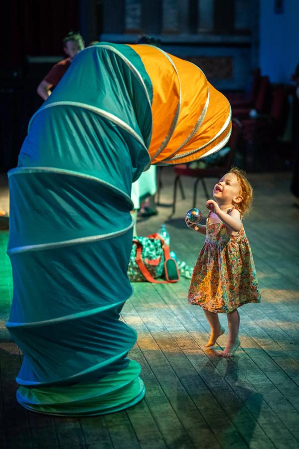 Child looks up to an actor on stage hidden inside a colourful fabric tube.