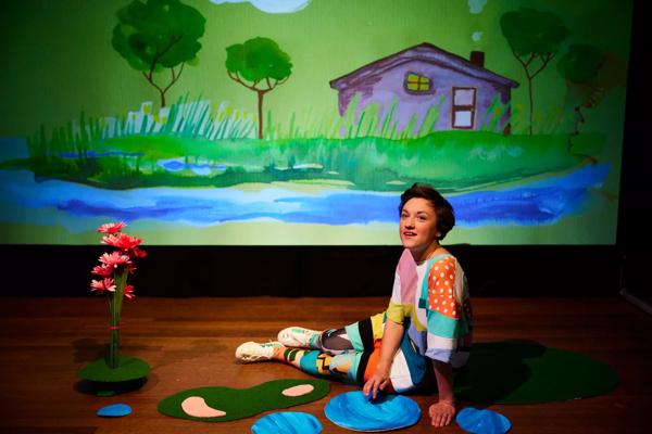 A performer sits on the floor with a patch of flowers in front of a cartoon backdrop showing trees and a house. 