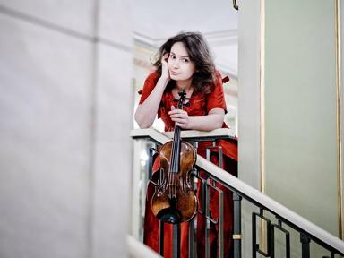 Woman stands on a staircase holding a violin, she is wearing a red dress. 