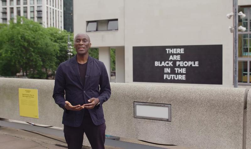 Curator Ekow Eshun stands outdoors at the Southbank Centre, behind him on the exterior wall of the Royal Festival Hall is Alisha B Wormsley's artwork 'There Are Black People in the Future'