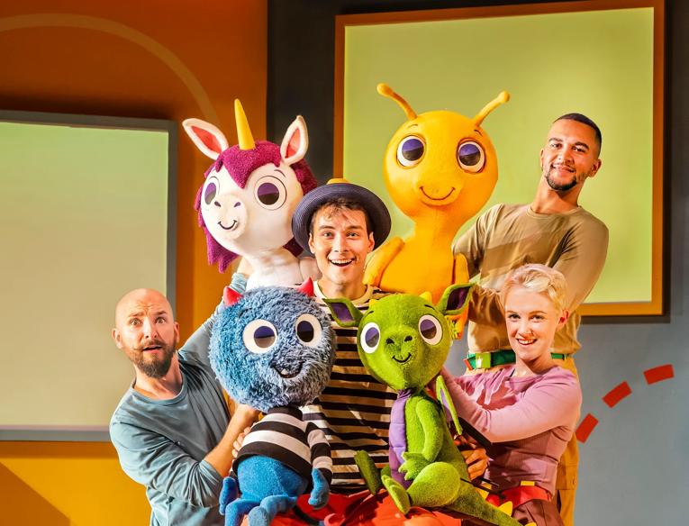 Four actors pose together holding a colourful monster character puppet.