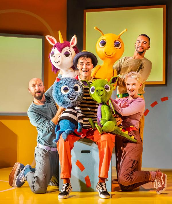 Four actors pose together holding a colourful monster character puppet.