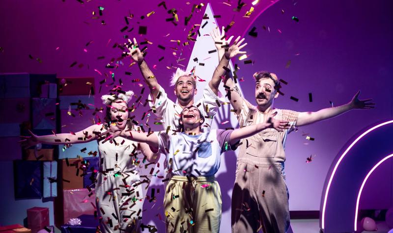 4 performers hold their arms up in celebration with smiles on their faces surrounded by floating metallic confetti.