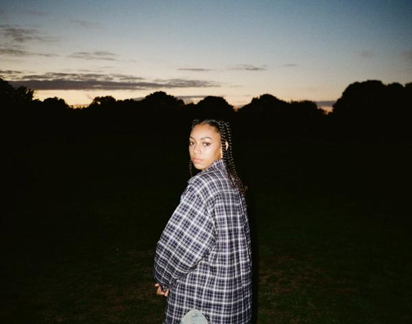 Person with braids, wearing a grey plaid oversized shirt standing in front of a sunset