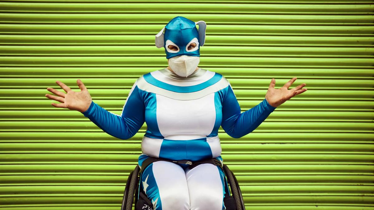 Comedian Jess Thom sits in her wheelchair in a metallic blue and white all in one suit with a matching mask covering the top half of her head. Her arms are spread wide and she wears a N95 mask over her nose and mouth. Behind her are bright green garage doors.