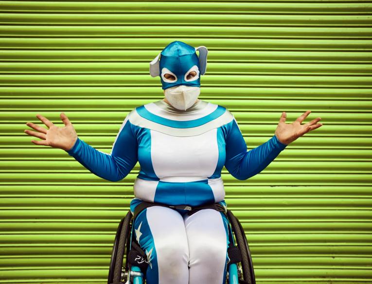 Comedian Jess Thom sits in her wheelchair in a metallic blue and white all in one suit with a matching mask covering the top half of her head. Her arms are spread wide and she wears a N95 mask over her nose and mouth. Behind her are bright green garage doors.