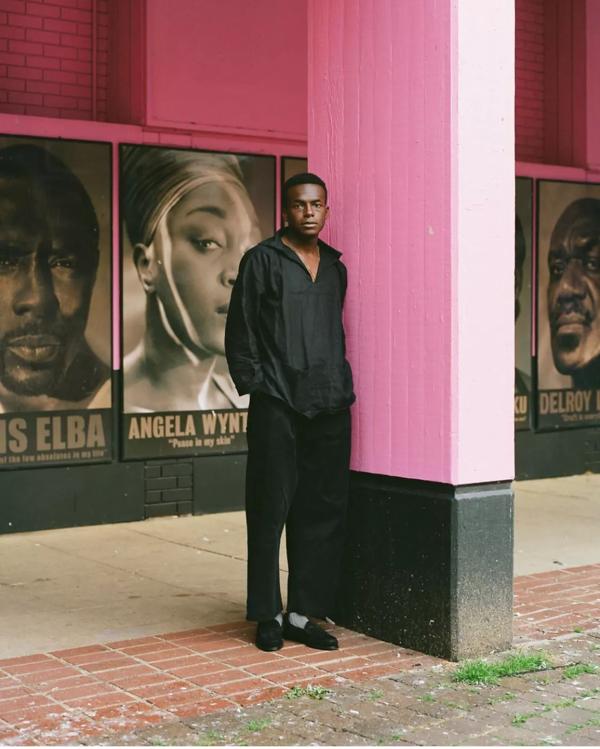 A Black man stands in all black against a pink column. 