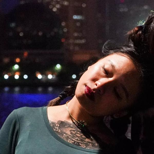 Poet Tim Tim Cheng has her head to one side, eyes closed, leaning against a wall. She has long dark hair, red lipstick and is wearing a green top, exposing some of her elaborate tattoo and a necklace. In the background is a city scene at night with a river reflecting high rise buildings.