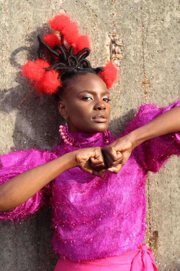 Portrait of singer and musician Shingai. She stands with her fists pushed together in front of her chest and wears a bright pink top and red fluffy pom poms in her hair.
