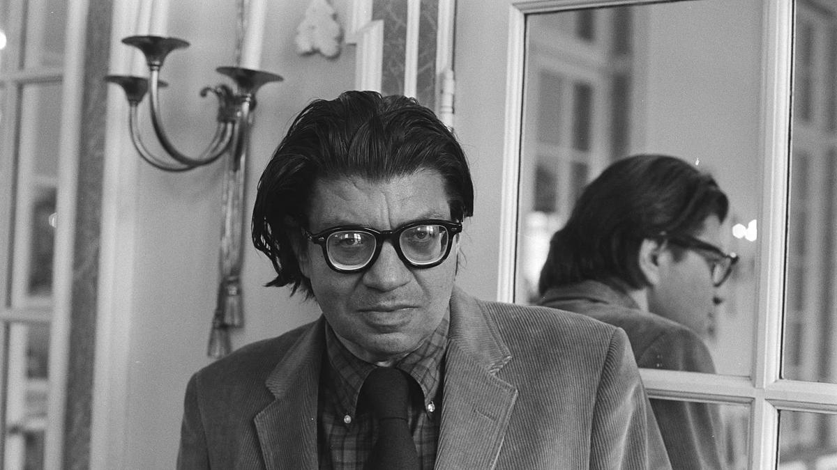 Composer Morton Feldman in black and white standing next to a mirrored door