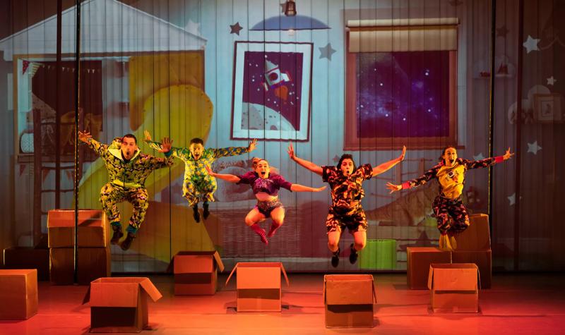 Five actors wearing pyjamas jumping over cardboard boxes.