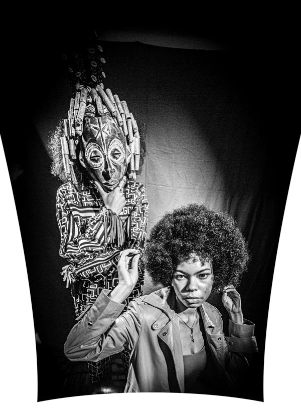 A black and white image featuring two Black woman, one of whom wears a traditional African mask