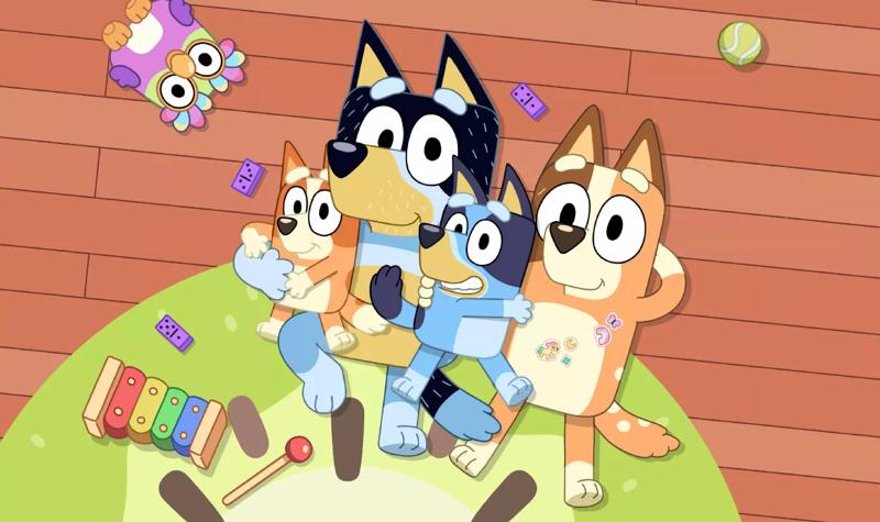 A cartoon dog family, two of which are blue and the other two are brown, lie on a green rug against a wooden floor gazing up at the sky. There is a pink owl toy, a ball and a xylophone on the rug with them.